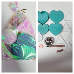 May include: A set of teal heart-shaped and triangular resin molds for crafting. The molds are packaged in a clear iridescent bag with a gold ribbon. The set also includes a small container of colorful rhinestones, a pink pen, and a gold glitter pen.