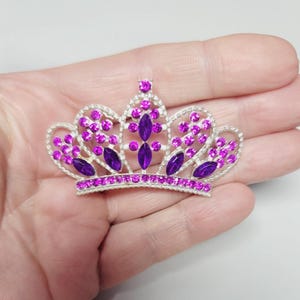 Peut inclure: Une couronne argentée ornée de pierres précieuses fuchsia et violettes. La couronne présente un design délicat avec de multiples arches et est ornée de cristaux scintillants et de gemmes en forme de goutte. Un petit bijou rond surmonte la couronne.