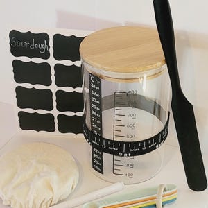 May include: A set of baking supplies including a glass jar with a wooden lid and a black spatula. The jar has markings for measuring in both Celsius and Fahrenheit. The jar also has a black tape measure around the middle with markings for measuring in milliliters and ounces. The jar is sitting on a white surface with a white cloth, a white marker, and a set of colorful measuring spoons.