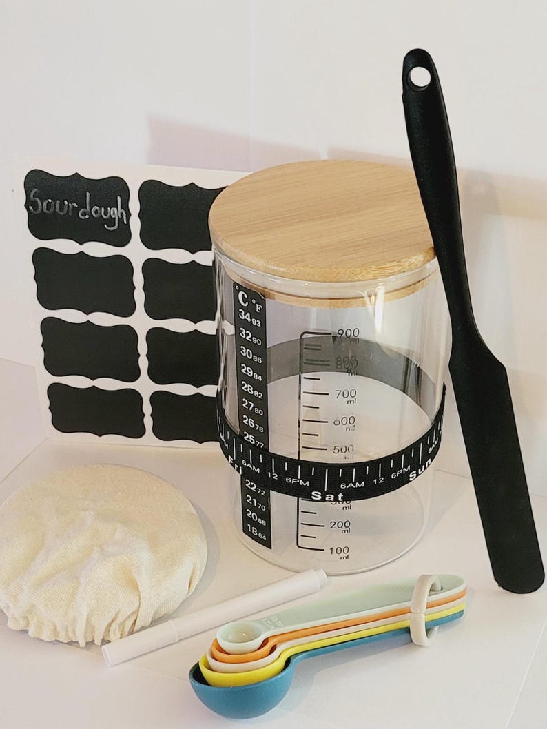 May include: A set of baking supplies including a glass jar with a wooden lid and a black spatula. The jar has markings for measuring in both Celsius and Fahrenheit. The jar also has a black tape measure around the middle with markings for measuring in milliliters and ounces. The jar is sitting on a white surface with a white cloth, a white marker, and a set of colorful measuring spoons.