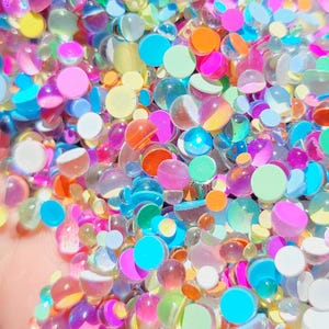May include: A close-up of a pile of colorful, translucent water beads. The beads are in various sizes and colors, including pink, blue, orange, yellow, and green. Some beads are solid, while others are clear with a colored center. The overall effect is vibrant and playful.
