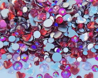 May include: A large assortment of red, pink, and clear rhinestones in various shapes, including round, heart, and flatback. The rhinestones are scattered on a white surface.