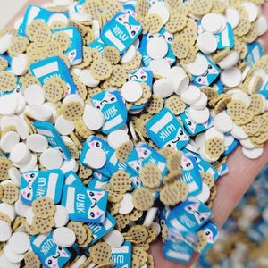 Op de afbeelding: Een close-up van een verscheidenheid aan kleurrijke sprinkles, waaronder blauwe melkpakken met een kattengezicht, bruine koekvormen en witte cirkels. De sprinkles zijn perfect om taarten, cupcakes en andere desserts te versieren.