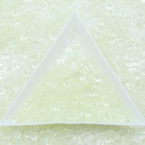 May include: A close-up of a white triangular tray filled with small, clear, round, iridescent rhinestones. The rhinestones are scattered across the tray and the background is a white surface.