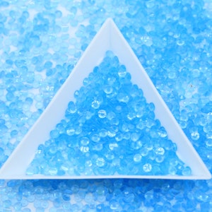 May include: A white triangular tray filled with light blue iridescent round beads. The tray is sitting on a white surface surrounded by more light blue beads.