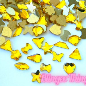 May include: Assortment of yellow rhinestone embellishments in various shapes, including stars, hearts, flowers, and geometric shapes. The rhinestones are scattered on a white background.