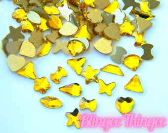 May include: Assortment of yellow rhinestone embellishments in various shapes, including stars, hearts, flowers, and geometric shapes. The rhinestones are scattered on a white background.