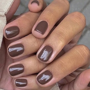 Puede incluir: Primer plano de una mano con uñas pintadas. Las uñas están pintadas de un color marrón oscuro brillante. La manicura está bien hecha, con un acabado suave y uniforme. El tono de piel es marrón claro y las uñas están bien formadas.