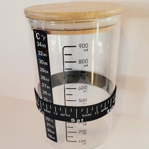 May include: A clear glass jar with a wooden lid and a black measuring tape around the middle. The jar has markings for both Celsius and Fahrenheit temperatures, as well as milliliters. The tape has markings for hours and days of the week.