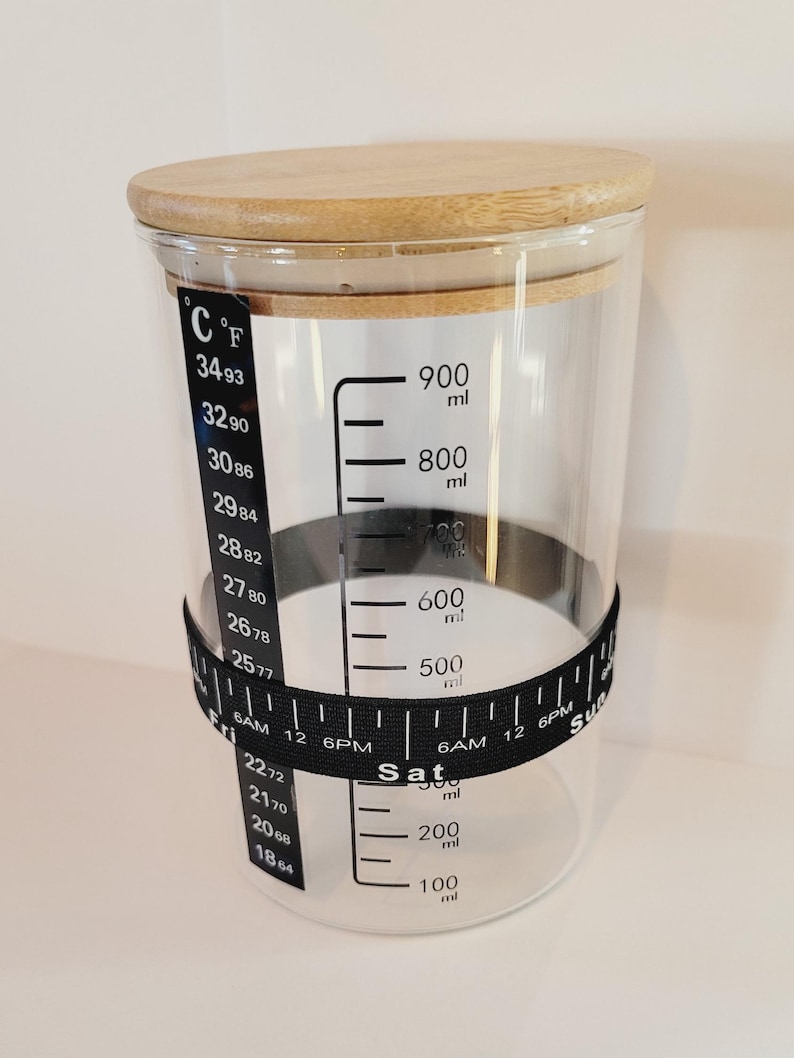 May include: A clear glass jar with a wooden lid and a black measuring tape around the middle. The jar has markings for both Celsius and Fahrenheit temperatures, as well as milliliters. The tape has markings for hours and days of the week.
