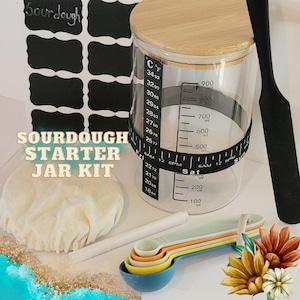 May include: A sourdough starter jar kit with a clear glass jar, a wooden lid, a white cloth, a black spatula, and measuring spoons. The jar has a black and white tape measure around it with markings for Celsius and Fahrenheit temperatures, as well as milliliters and ounces. The jar also has a black and white label that says "Sourdough Starter Jar Kit".