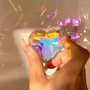 May include: A clear heart-shaped crystal prism with rainbow facets. The crystal is being held in a hand, and it is casting rainbows on the background.