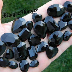 Peut inclure: Une collection de pierres précieuses en verre facetté noir de différentes formes, notamment des ovales, des cœurs et des larmes. Les pierres précieuses sont disposées sur une main, avec le texte "Blingee Thingie" visible en arrière-plan.