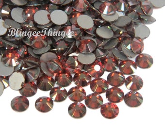May include: A collection of faceted, round, dark red rhinestones with a metallic silver backing. The rhinestones are scattered across a white surface, reflecting light. The text "BlingeeThings" is visible in the upper left corner.