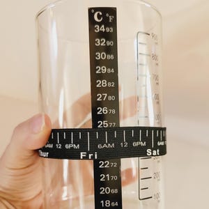 May include: A clear glass measuring cup with black markings for both Celsius and Fahrenheit temperatures. A black elastic band with a ruler and day of the week markings is wrapped around the cup.