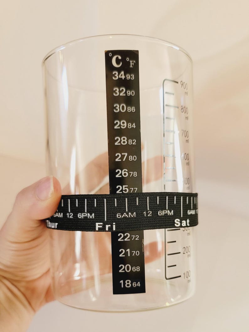 May include: A clear glass measuring cup with black markings for both Celsius and Fahrenheit temperatures. A black elastic band with a ruler and day of the week markings is wrapped around the cup.