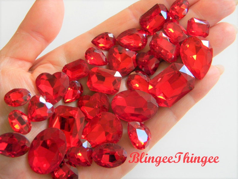 May include: A collection of red glass rhinestones in various shapes and sizes, including round, oval, teardrop, and heart shapes. The rhinestones are scattered on a hand, with the text "BlinggeeThingee" visible in the bottom right corner.