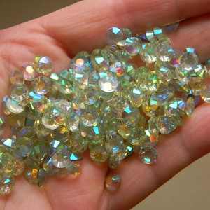 Puede incluir: Un montón de piedras de cristal verdes y transparentes iridiscentes en una mano. Las piedras de cristal son facetadas y tienen un brillo arcoíris.