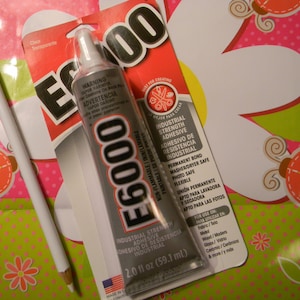 May include: A tube of E6000 industrial strength adhesive, 2.0 fl oz, with a white pencil on a pink and green floral patterned background. The adhesive is non-flammable and suitable for fabric, wood, glass, and ceramics.