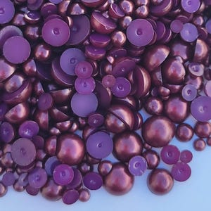 May include: An overhead shot of a pile of purple and maroon beads. The beads are various sizes and shapes, including whole spheres and half-spheres. Some beads have a glossy finish, while others are matte. The beads are scattered across a white surface.