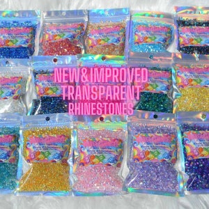 Pode incluir: Várias sacolas plásticas transparentes cheias de strass de resina coloridos. As sacolas estão dispostas em fileiras e colunas. O texto "NEW & IMPROVED TRANSPARENT RHINESTONES" é exibido em rosa no centro da imagem.