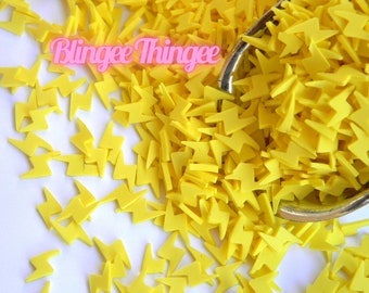 May include: A close-up of a yellow lightning bolt sprinkles. The sprinkles are in a small silver scoop and scattered on a white surface. The text "Blinggee Thinggee" is visible in pink letters.