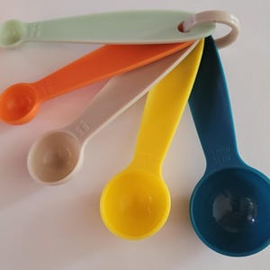 May include: A set of five colorful measuring spoons with a ring handle. The spoons are in shades of blue, yellow, beige, orange, and green. The spoons are labeled with measurements in tablespoons and milliliters.