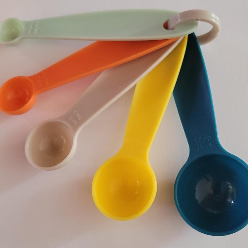 May include: A set of five colorful measuring spoons with a ring handle. The spoons are in shades of blue, yellow, beige, orange, and green. The spoons are labeled with measurements in tablespoons and milliliters.
