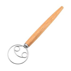 Puede incluir: Un batidor de masa danés con un mango de madera de color marrón claro y un lazo de alambre de acero inoxidable. El batidor está diseñado para mezclar masa y batidos. El mango de madera se estrecha hasta un extremo redondeado.