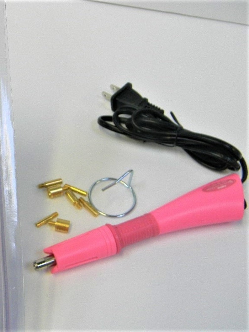 Puede incluir: Una herramienta aplicadora de pedrer&iacute;a rosa con un cable de alimentaci&oacute;n negro, un anillo de metal plateado y varias puntas de pedrer&iacute;a doradas. La herramienta tiene un mango rosa y un agarre rojo.