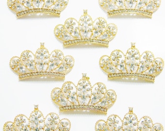 Pode incluir: Oito coroas douradas com detalhes em strass transparente. As coroas estão dispostas simetricamente sobre um fundo branco.