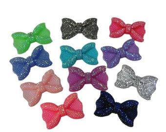 May include: Twelve sparkly resin bows in various colors, including pink, blue, green, purple, and silver. Each bow has a slightly different shape and size.
