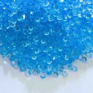 May include: A pile of small, light blue, faceted, round, glass gems. The gems are scattered on a white surface.