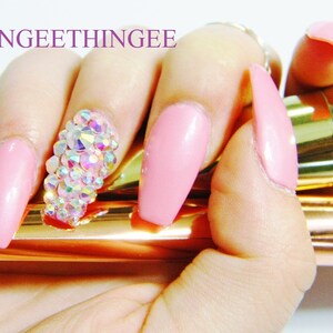 May include: Pink long acrylic nails with a large cluster of iridescent rhinestones on one nail. The nails are held by a hand holding two gold makeup brushes.