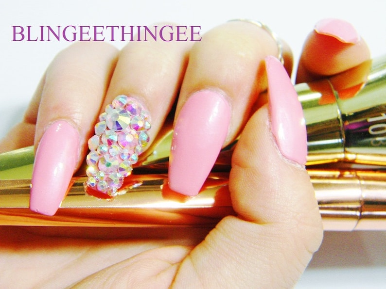 May include: Pink long acrylic nails with a large cluster of iridescent rhinestones on one nail. The nails are held by a hand holding two gold makeup brushes.