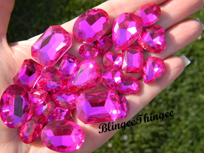 May include: A collection of pink, faceted, glass rhinestones of various shapes and sizes. The rhinestones are arranged in a hand, with the text "Blingyee Thinghee" visible in the lower right corner.