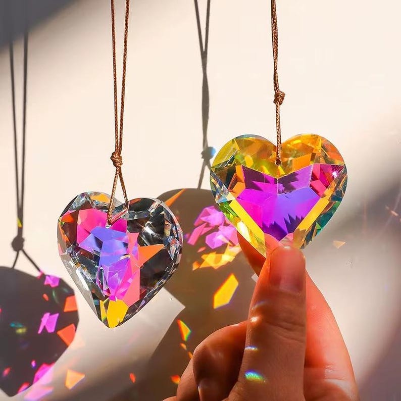 May include: Two iridescent heart-shaped crystal prisms hanging from brown cords. The prisms are catching the light and creating rainbows.