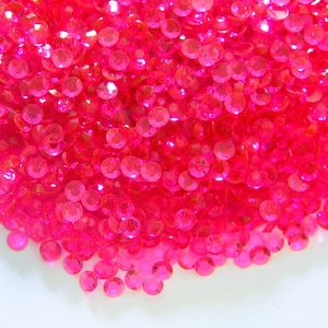 May include: A close-up shot of a pile of small, round, hot pink rhinestones. The rhinestones are scattered and reflect light, creating a sparkling effect. The image is brightly lit, highlighting the color and texture of the rhinestones.