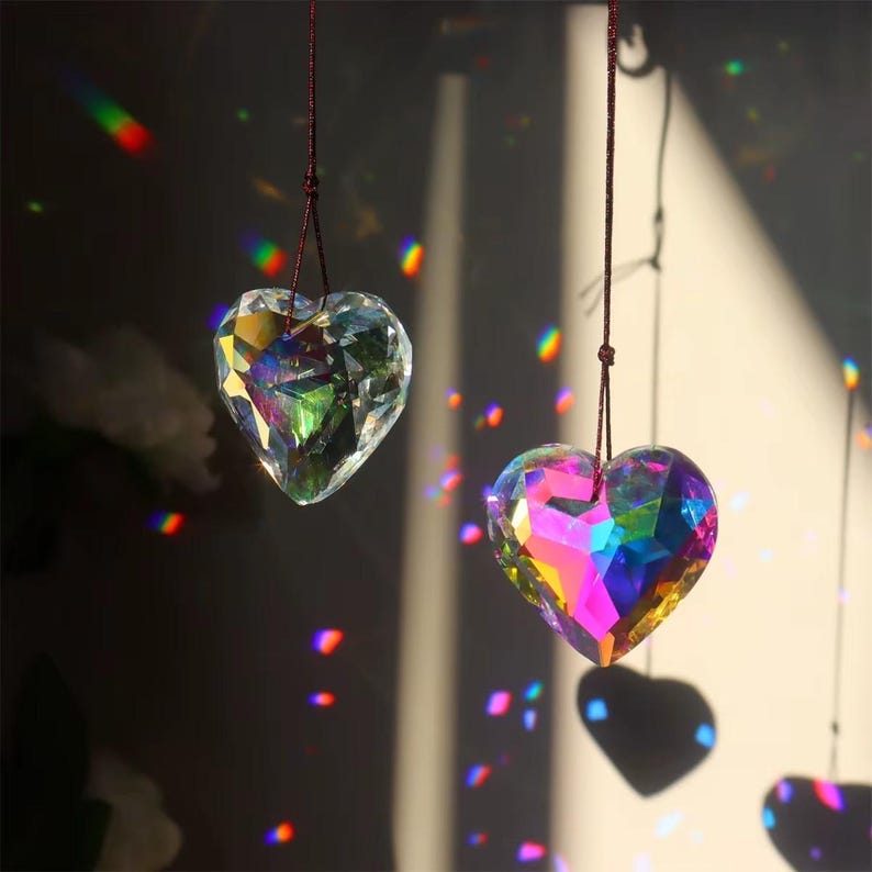 May include: Two iridescent heart-shaped crystal prisms hanging from strings. The prisms are clear and reflect rainbow colors.