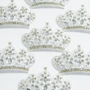 May include: Several silver-toned crowns with clear, sparkling rhinestones. Each crown has a decorative band at the base and is embellished with various sizes of rhinestones. The crowns are arranged on a white background.