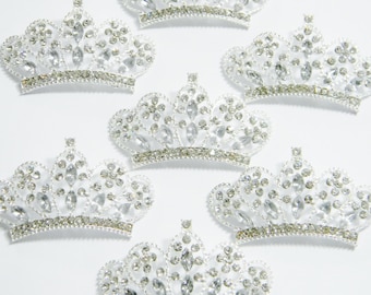 Pode incluir: Várias coroas prateadas com strass transparentes e brilhantes. Cada coroa tem uma faixa decorativa na base e é adornada com strass brilhantes de vários tamanhos. As coroas estão dispostas sobre um fundo branco.