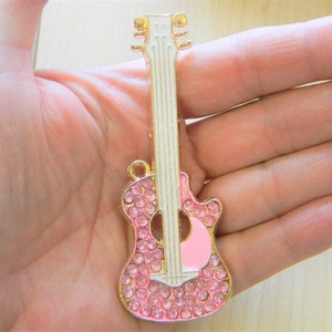 Könnte beinhalten: Ein rosa und goldener Gitarrenanhänger mit Strasssteinen. Die Gitarre hat einen weißen Korpus und einen goldenen Hals.