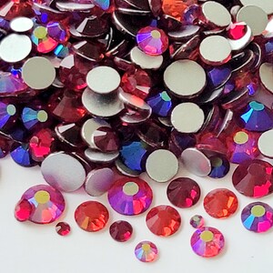 May include: A large pile of red, purple, and blue iridescent rhinestones. The rhinestones are scattered on a white surface. Some of the rhinestones have flat backs, while others have pointed backs.
