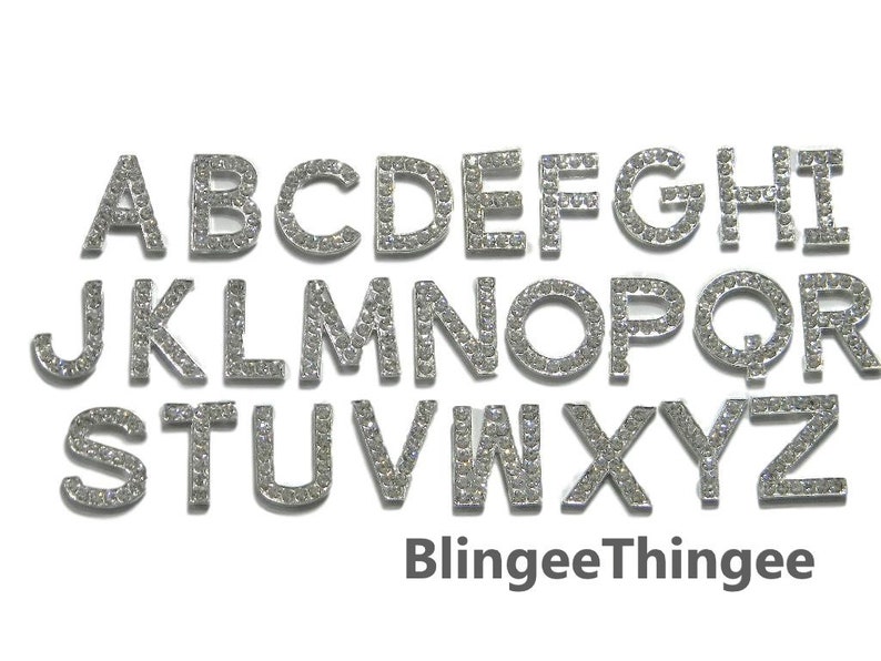 Pode incluir: Um conjunto de 26 letras prateadas, cada uma incrustada com strass transparentes, soletrando o alfabeto de A a Z. As letras est&atilde;o dispostas em tr&ecirc;s linhas. O texto "BlingeeThingee" est&aacute; impresso abaixo das letras.