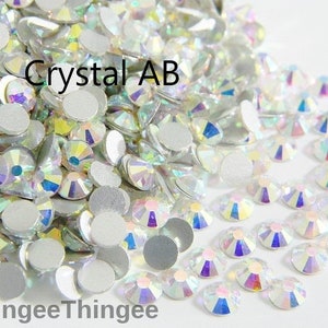 May include: A close-up image of a large quantity of clear crystal AB rhinestones. The rhinestones are scattered on a white surface. The text "Crystal AB" is visible in the image.