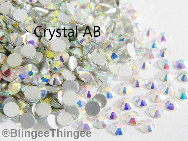 May include: A close-up image of a large quantity of clear crystal AB rhinestones. The rhinestones are scattered on a white surface. The text "Crystal AB" is visible in the image.