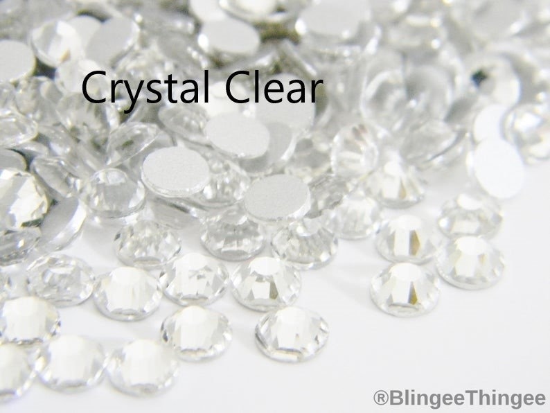 May include: A close-up image of a pile of clear crystal rhinestones. The rhinestones are small and have a faceted design. The text "Crystal Clear" is visible in the image.