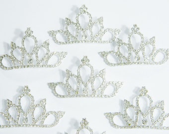 Pode incluir: Seis coroas prateadas com detalhes em strass transparentes. As coroas estão dispostas de forma dispersa sobre um fundo branco.