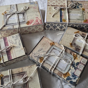 Könnte beinhalten: Mehrere Geschenkboxen im Vintage-Stil mit Designs aus gealtertem Papier, floralen Akzenten und Bindfäden. Die Boxen haben gerahmte Etiketten mit dem Text "All we need..." und andere handschriftliche Details. Die Gesamtästhetik ist rustikal und nostalgisch.