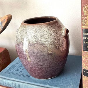May include: A rounded ceramic vase featuring a two-tone glaze with a dark purple base that transitions into a speckled cream-colored top. The vessel has a wide opening and small, integrated side handles, showcasing a smooth, glossy finish.
